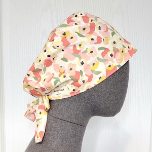 Peut inclure: Un bonnet de chirurgien à motifs floraux dans les tons de rose, de jaune et de vert sur fond crème. Le bonnet est noué dans le dos avec des liens en tissu assortis. L'étiquette indique "Mombi Calok".