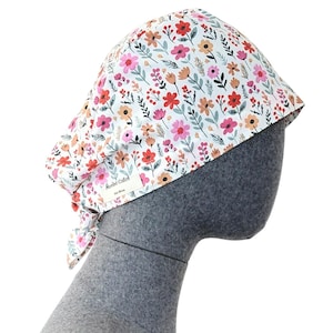 Peut inclure: Un foulard à imprimé floral avec un fond blanc orné de fleurs roses, rouges et oranges. Le foulard est noué dans le dos et porte une petite étiquette "Monbo Catco".