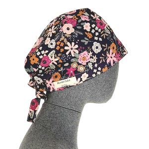 Peut inclure: Une toque de chirurgien à motif floral, avec un fond bleu marine foncé et des fleurs roses, blanches et orange. Le bonnet a une attache à l'arrière et une petite étiquette "Mombó Calot".