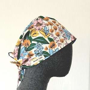 Peut inclure: Une charlotte de bloc opératoire à fond blanc ornée d'un motif floral coloré dans des tons rose, jaune, bleu clair et vert foncé. Le tissu est noué à l'arrière et comporte une petite étiquette rectangulaire blanche avec le texte Mombô Calot.