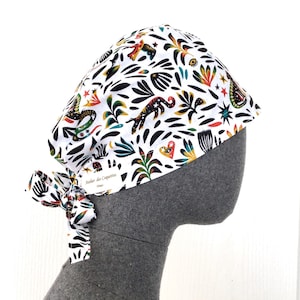 Peut inclure: Un bonnet de chirurgien blanc avec un motif coloré d'animaux stylisés, de plantes et d'étoiles. Le bonnet a une attache à l'arrière et le texte "Atelier des Coquettes" est imprimé sur l'attache.