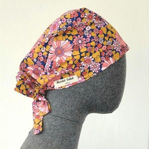 Peut inclure: Une charlotte de bloc opératoire avec un motif floral aux tons rose, jaune moutarde et bleu marine. Elle possède un lien à nouer à l'arrière et une petite étiquette rectangulaire blanche sur le côté portant le texte noir Mombö Calot Nîmes.