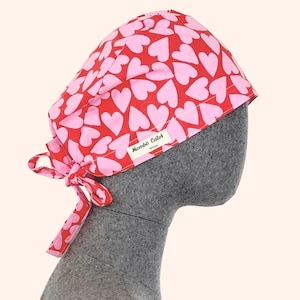 Peut inclure: Une charlotte de bloc rouge avec un motif répétitif de cœurs rose clair. Le bonnet possède un lien de serrage à l'arrière et une petite étiquette rectangulaire blanche sur le côté avec le texte noir Mombô Calot Nîmes imprimé.