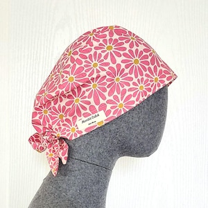 Peut inclure: Un couvre-chef à motifs floraux roses et jaunes avec un nœud. Le couvre-chef est sur une tête de mannequin grise. Le texte "Mombô Color" est sur une petite étiquette.