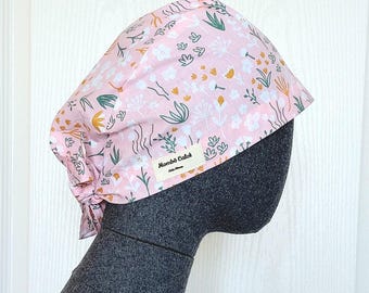 May include: A pink floral patterned scrub cap with white, yellow, and green accents. The cap is tied at the back and features a small label that reads "Mombô Caloi". The cap is displayed on a gray mannequin head.