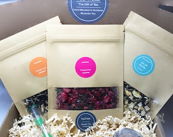 May include: Three brown paper bags filled with loose leaf tea blends. The bags are labelled "Ginger Nut", "Pretty in Pink", and "Get Up And Go". The bags are in a box with shredded paper and a tea strainer.