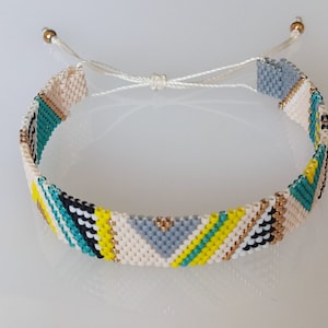 Peut inclure: Un bracelet en perles coloré avec un motif géométrique dans des tons de blanc, gris, noir, jaune et turquoise. Le bracelet a un cordon blanc avec des perles dorées aux extrémités.
