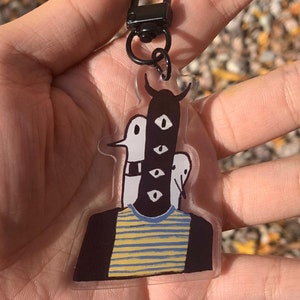 Peut inclure: Un porte-cl&eacute;s en acrylique transparent avec une illustration fantaisiste d'une figure &agrave; plusieurs yeux et une t&ecirc;te cornue. La figure porte un t-shirt ray&eacute; bleu et jaune et est attach&eacute;e &agrave; un fermoir en m&eacute;tal noir.