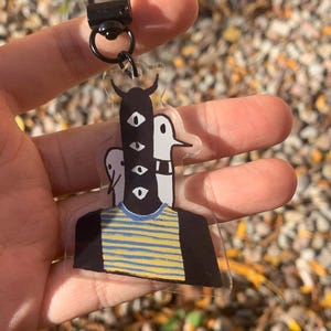 Peut inclure: Un porte-cl&eacute;s en acrylique transparent avec une illustration fantaisiste d'une figure avec plusieurs yeux et une chemise ray&eacute;e. Le porte-cl&eacute;s a un fermoir et un anneau noirs. L'arri&egrave;re-plan est flou avec des feuilles d'automne.