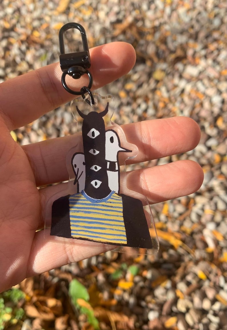 Peut inclure: Un porte-cl&eacute;s en acrylique transparent avec une illustration fantaisiste d'une figure avec plusieurs yeux et une chemise ray&eacute;e. Le porte-cl&eacute;s a un fermoir et un anneau noirs. L'arri&egrave;re-plan est flou avec des feuilles d'automne.
