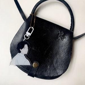 May include: A black leather satchel bag with a top handle and a shoulder strap. A clear acrylic keychain with a cartoon illustration of a person is attached to the bag.