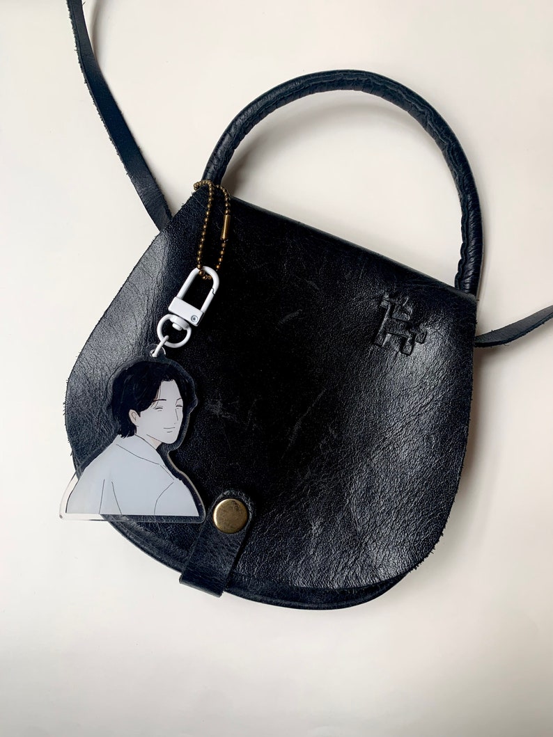 May include: A black leather satchel bag with a top handle and a shoulder strap. A clear acrylic keychain with a cartoon illustration of a person is attached to the bag.