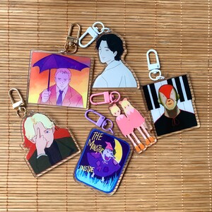 May include: Six colorful keychains featuring various characters and designs. The keychains are made of clear acrylic and have metal rings for attaching to keys or bags. The designs include a man with an umbrella, a man with a red and black design, two women with pink dresses, a man with a black and white design, and a man with a black and white design.