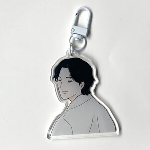 May include: A white keychain with a clear acrylic charm featuring a black and white line drawing of a man with black hair. The man is smiling and looking to the side.
