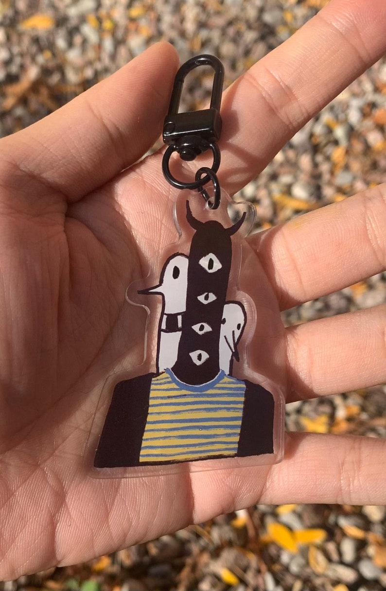 Peut inclure: Un porte-cl&eacute;s en acrylique transparent avec une illustration fantaisiste d'une figure &agrave; plusieurs yeux et une t&ecirc;te cornue. La figure porte un t-shirt ray&eacute; bleu et jaune et est attach&eacute;e &agrave; un fermoir en m&eacute;tal noir.