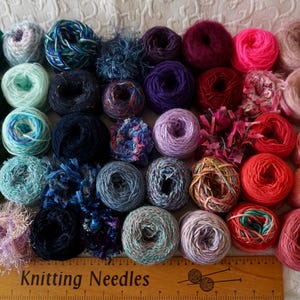 May include: A selection of colourful yarn balls arranged in a gradient, from greens and blues to pinks and yellows. A wooden ruler with the text "Knitting Needles" is visible. These yarn balls are suitable for knitting and other craft projects.