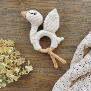 May include: A white crocheted swan rattle with a wooden ring and tan crocheted tail. The rattle is on a wooden surface.