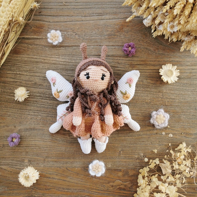 May include: A handmade crochet fairy doll with brown hair, a pink dress, and white wings decorated with floral accents. The doll has a brown cap with antennae and white shoes. Decorative flowers surround the doll on a wooden surface.