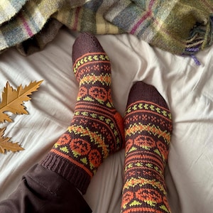 Puede incluir: Un par de calcetines de punto marrón con un estampado colorido de otoño con calabazas, hojas y formas geométricas. Los calcetines están puestos en los pies de una persona.