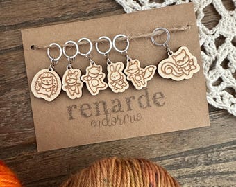 May include: A set of six wooden stitch markers featuring woodland animal designs. The markers are attached to silver rings and are displayed on a brown card with the text "renarde endormie".