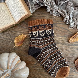 Peut inclure: Une paire de chaussettes en tricot marron et crème avec un motif d'écusson. Les chaussettes sont sur une surface en bois avec un livre, une couverture et des feuilles d'automne.