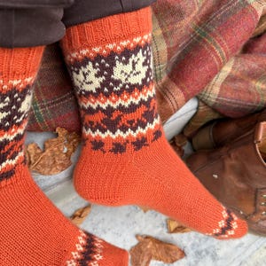 May include: A pair of orange knit socks with a geometric pattern in cream and brown. The socks are visible from the ankle down, showcasing the detailed design. The socks are resting on a surface with fallen leaves and a plaid blanket.