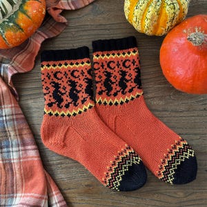 Puede incluir: Un par de calcetines de punto naranja con talones y dedos negros. Los calcetines presentan un patrón de siluetas de brujas negras, lunas y estrellas, con detalles en amarillo y negro. Los calcetines se muestran con calabazas y una tela a cuadros.