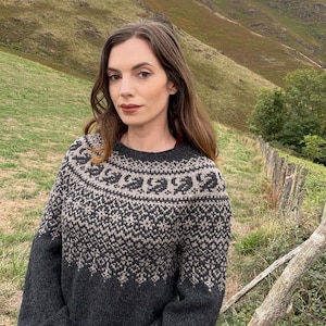 Könnte beinhalten: Ein langärmeliger Strickpullover mit einer dunkel anthrazitfarbenen Basis und einer kontrastierenden hellbeigen Passe. Die Passe zeigt ein detailliertes kreisförmiges Muster aus geometrischen Formen und stilisierten Blattmotiven.