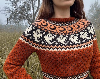 May include: A crew neck sweater in shades of orange, black, and cream. The sweater features a patterned design across the chest and sleeves. The background is a field of tall grass and a tree.