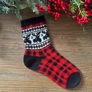 Peut inclure: Une chaussette en tricot noire et rouge avec un motif blanc et noir. La chaussette a un motif de plaid buffalo rouge et noir sur le pied et un motif blanc et noir avec un motif d'arbre de Noël sur la jambe.