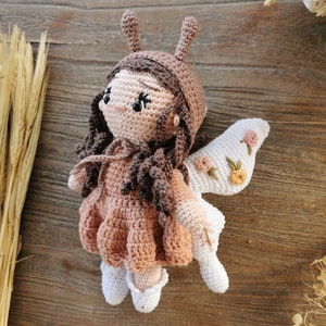 May include: A handmade crochet doll with a fairy theme. The doll has a light brown skin tone, brown hair, and a pink dress. It features white wings with embroidered flowers. The doll is approximately 8 inches tall.