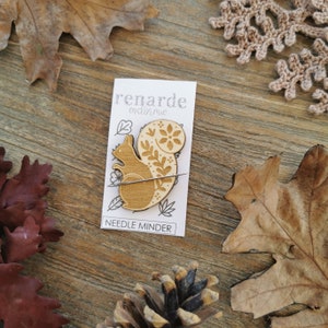 May include: Wooden needle minder shaped like a squirrel with a floral design. The needle minder is on a white card with the text "renarde endormie" and "NEEDLE MINDER".