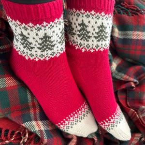 May include: A pair of red knitted socks with a white and green Christmas tree pattern. The socks are on a plaid blanket.