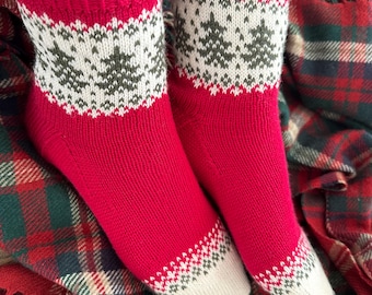 May include: A pair of red knitted socks with a white and green Christmas tree pattern. The socks are on a plaid blanket.