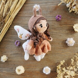 May include: A handmade crochet fairy doll with brown hair, a tan dress, and white wings with floral embroidery. The doll has a brown hood with small ears and is surrounded by decorative flowers on a wooden surface.