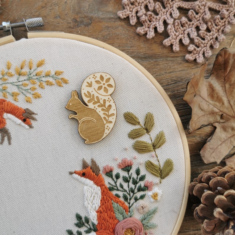 May include: A wooden squirrel with a floral design on its tail, sitting on a white fabric background with embroidered flowers and leaves. The squirrel is a light brown colour and the flowers are pink, white, and green.
