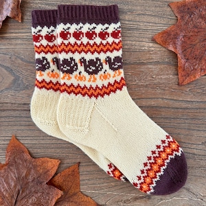 May include: A pair of cream-coloured knitted socks with a brown, orange and white patterned design featuring turkeys and apples. The socks have a brown cuff.