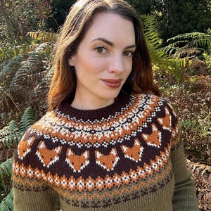 Peut inclure: Un pull tricoté vert, marron et orange, avec un col rond. Le pull présente un motif de renards et de formes géométriques en marron, blanc et orange. La personne porte le pull à l'extérieur.