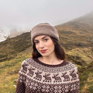 Peut inclure: Un pull en tricot marron et beige avec un motif de rennes. Le pull est associé à un chapeau marron duveteux assorti. La femme est à l'extérieur, devant une montagne.