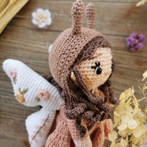 May include: A handmade crochet doll with a fairy design. The doll features a tan face, brown hair, and a brown bonnet with antennae. It has white wings with floral embroidery and is wearing a pink dress. The doll is a whimsical, handcrafted item.