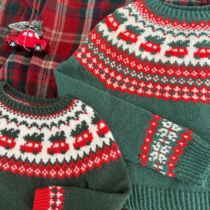 Puede incluir: Dos jerséis de punto verdes con un patrón festivo de diseños rojos y blancos. Los jerséis tienen cuello redondo y mangas largas. Un pequeño coche de juguete rojo con un árbol de Navidad está sobre un fondo de cuadros rojos.