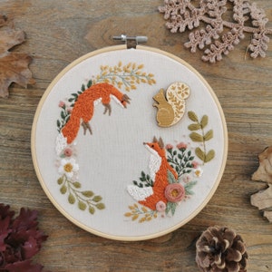 May include: A white embroidery hoop with a floral design featuring two foxes and a squirrel. The foxes are orange and the squirrel is brown. The hoop is on a wooden surface.