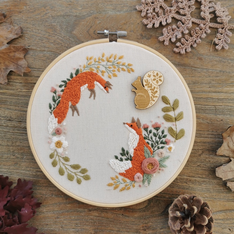 May include: A white embroidery hoop with a floral design featuring two foxes and a squirrel. The foxes are orange and the squirrel is brown. The hoop is on a wooden surface.
