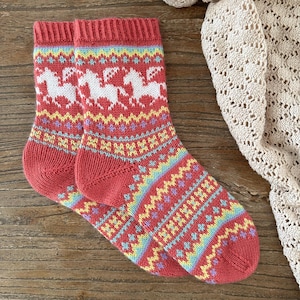 Op de afbeelding: Een paar rode gebreide sokken met een wit paardmotief en kleurrijke geometrische patronen.