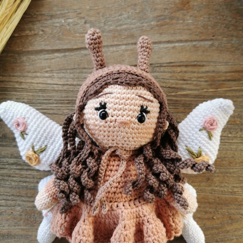 May include: A handmade crochet fairy doll with brown hair, antennae, and a pink dress. The doll has white wings with embroidered pink and yellow flowers. The doll is made of soft yarn and is a whimsical toy.