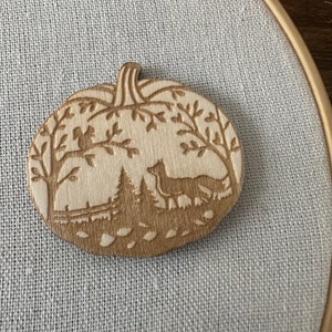 May include: A wooden pumpkin ornament with a carved design of a forest scene with trees, a fox, and birds. The ornament is painted in shades of brown and beige.