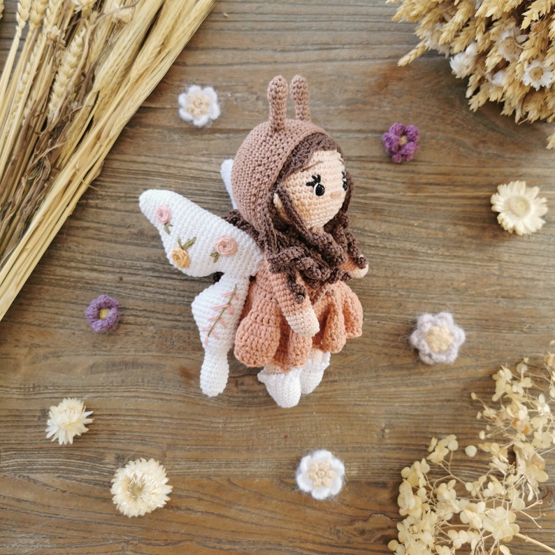 May include: A handmade crochet fairy doll with a brown hood and dress, white wings, and brown hair. The wings are decorated with embroidered flowers. The doll is surrounded by small crochet flowers and dried wheat on a wooden surface.