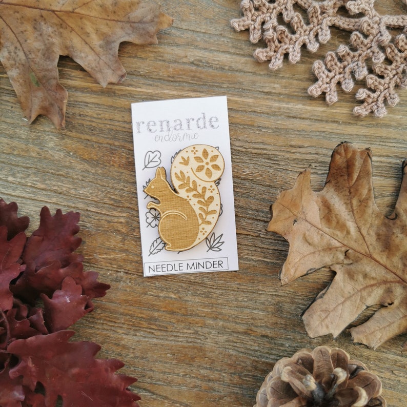 May include: Wooden needle minder featuring a squirrel with a floral design on its tail. The needle minder is on a white card with the text "remarde" and "needle minder".