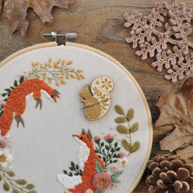 May include: A white embroidery hoop with a floral design featuring two foxes and a squirrel. The embroidery is done in shades of orange, brown, and green. The hoop is on a wooden surface.