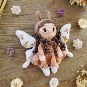 May include: A handmade crochet fairy doll with brown hair, a pink dress, and white wings adorned with embroidered flowers. The doll has a brown hood and is surrounded by small crochet flowers on a wooden surface.